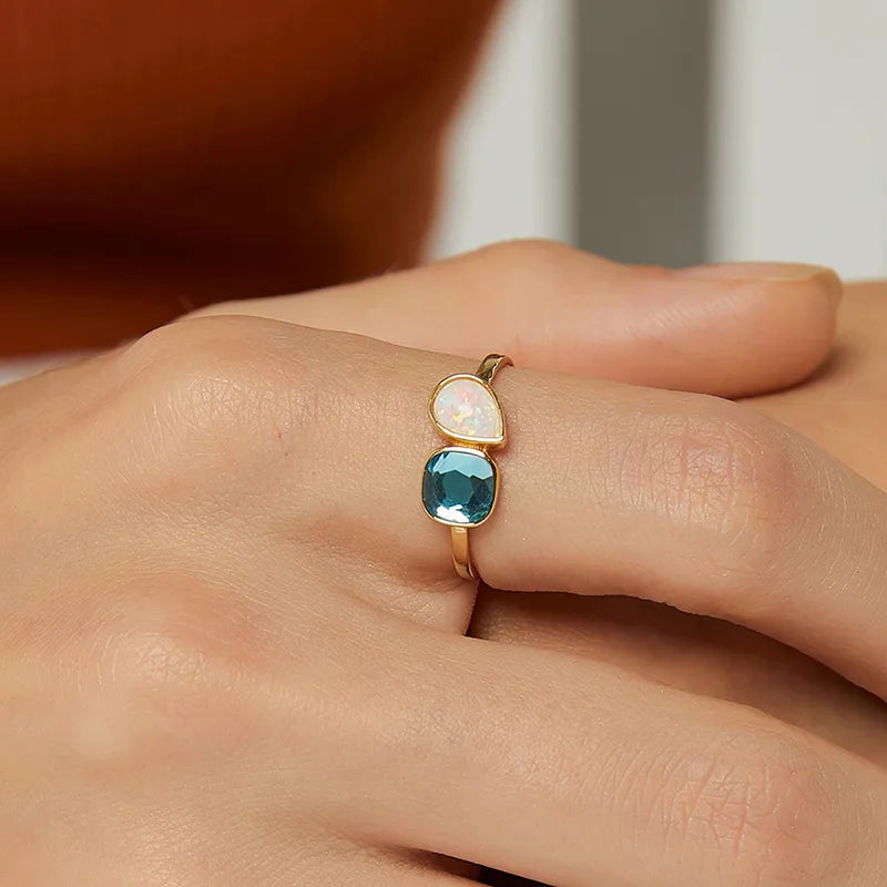 Bague dorée or 18K pour femme, ornée de deux pierres asymétriques – une bleue facettée et une blanche effet opale –, au design moderne et lumineux, parfaite pour un style estival, créatif et élégant.