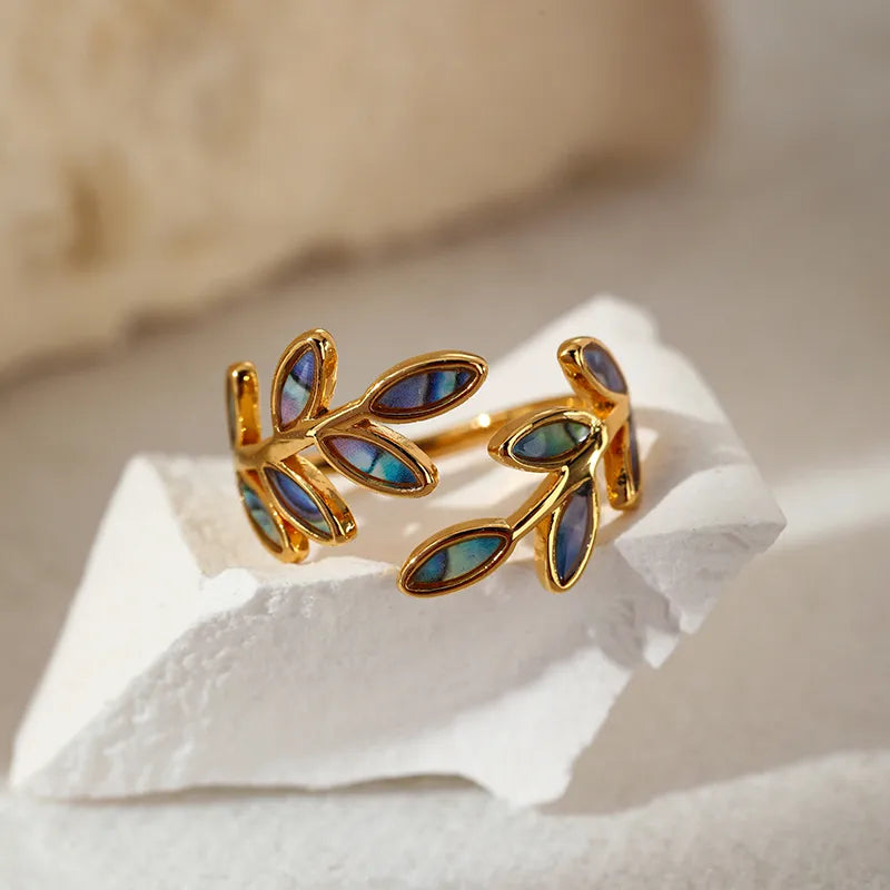 Apportez une touche d’élégance naturelle à votre style avec cette bague en acier inoxydable doré, ornée de délicates feuilles incrustées de coquille d’ormeau. Son design ouvert épouse parfaitement la forme du doigt tout en reflétant la lumière avec subtilité grâce aux reflets irisés de la nacre.