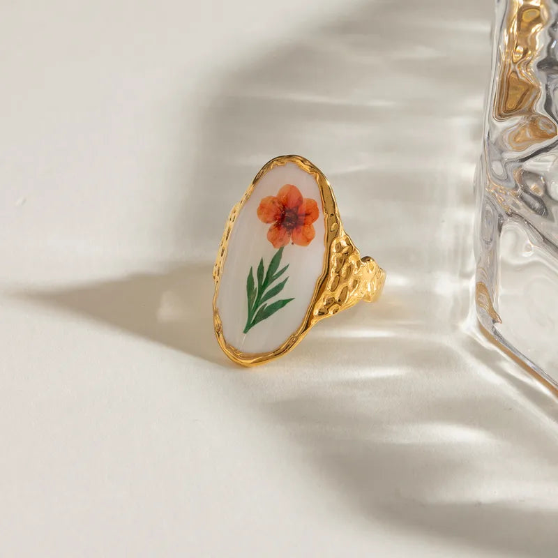 Laissez éclore votre style avec cette bague dorée en acier inoxydable au charme artistique. Ornée d’un médaillon ovale blanc illustré d’une fleur orange peinte à la main, cette bague florale vintage marie le romantisme d’un herbier à l’éclat du métal martelé.