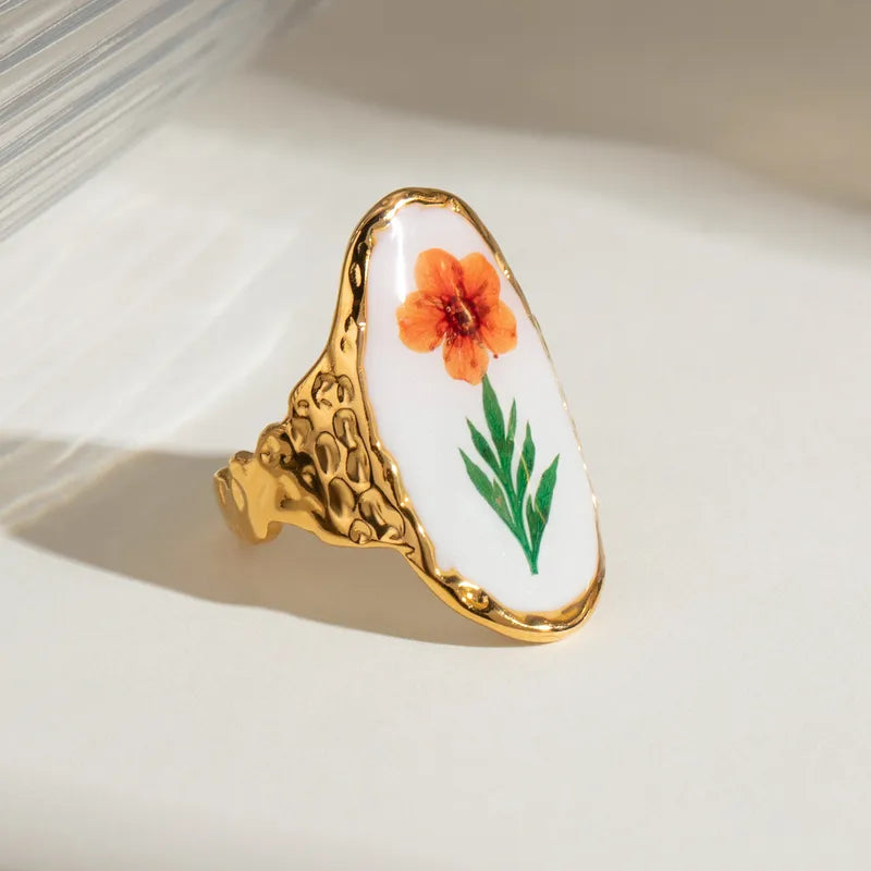 Laissez éclore votre style avec cette bague dorée en acier inoxydable au charme artistique. Ornée d’un médaillon ovale blanc illustré d’une fleur orange peinte à la main, cette bague florale vintage marie le romantisme d’un herbier à l’éclat du métal martelé.