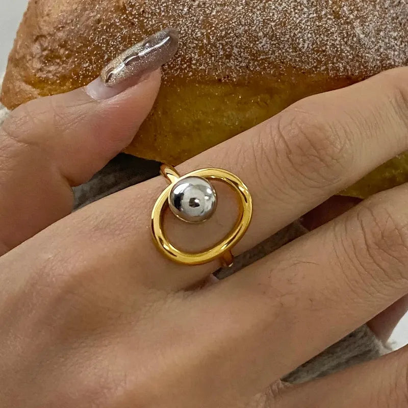 Bague en acier inoxydable doré au design géométrique moderne, ornée d’une sphère argentée brillante insérée dans un cercle ouvert, pour un style chic et contemporain.