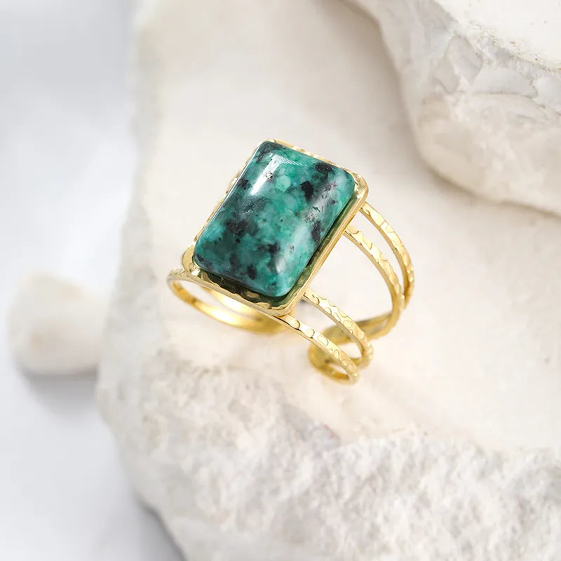 Affirmez une allure audacieuse avec cette bague dorée pour femme en acier inoxydable, à la fois robuste et élégante. Son design ajouré et martelé lui confère un look artisanal unique, tandis que sa grande pierre chrysocolle naturelle attire