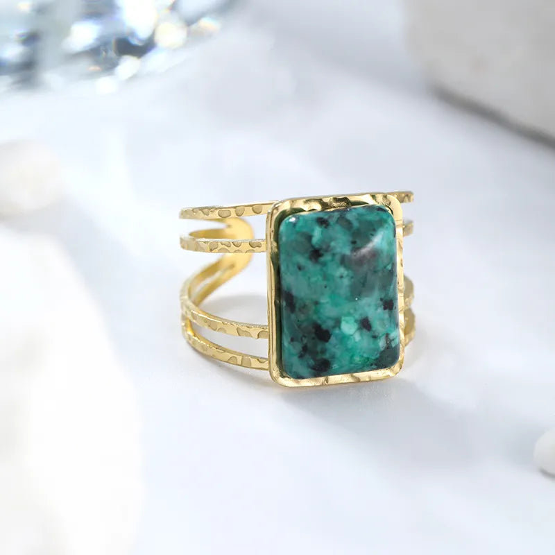 Affirmez une allure audacieuse avec cette bague dorée pour femme en acier inoxydable, à la fois robuste et élégante. Son design ajouré et martelé lui confère un look artisanal unique, tandis que sa grande pierre chrysocolle naturelle attire