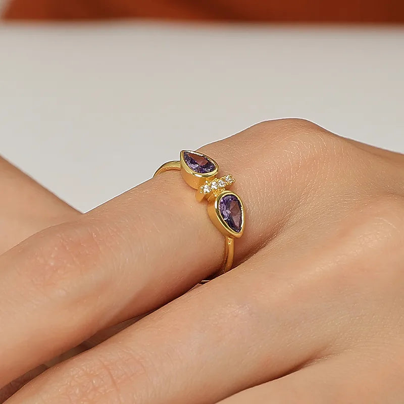 Bague dorée or 18K pour femme, ornée de deux pierres violettes en forme de goutte séparées par une ligne de zircons scintillants, au design ouvert asymétrique et raffiné, idéale pour un style romantique, coloré et sophistiqué.