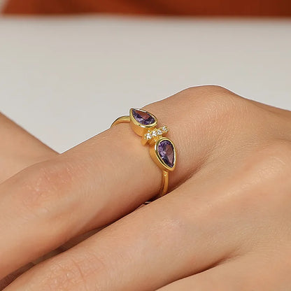 Bague dorée or 18K pour femme, ornée de deux pierres violettes en forme de goutte séparées par une ligne de zircons scintillants, au design ouvert asymétrique et raffiné, idéale pour un style romantique, coloré et sophistiqué.