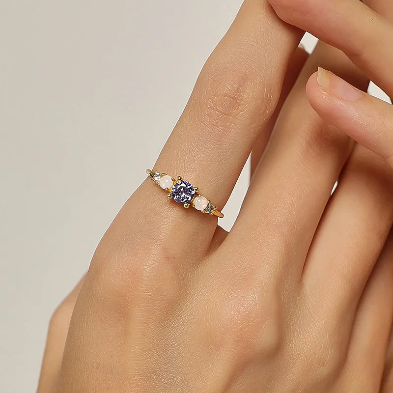 Cette bague féminine joue la carte du raffinement lumineux grâce à une composition délicate de pierres aux reflets changeants. Au centre, une pierre violette facettée attire tous les regards, accompagnée de deux opales blanches aux reflets irisés et de zircons scintillants de chaque côté. Un design équilibré, pensé pour capter la lumière et souligner la grâce de la main.