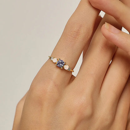 Cette bague féminine joue la carte du raffinement lumineux grâce à une composition délicate de pierres aux reflets changeants. Au centre, une pierre violette facettée attire tous les regards, accompagnée de deux opales blanches aux reflets irisés et de zircons scintillants de chaque côté. Un design équilibré, pensé pour capter la lumière et souligner la grâce de la main.