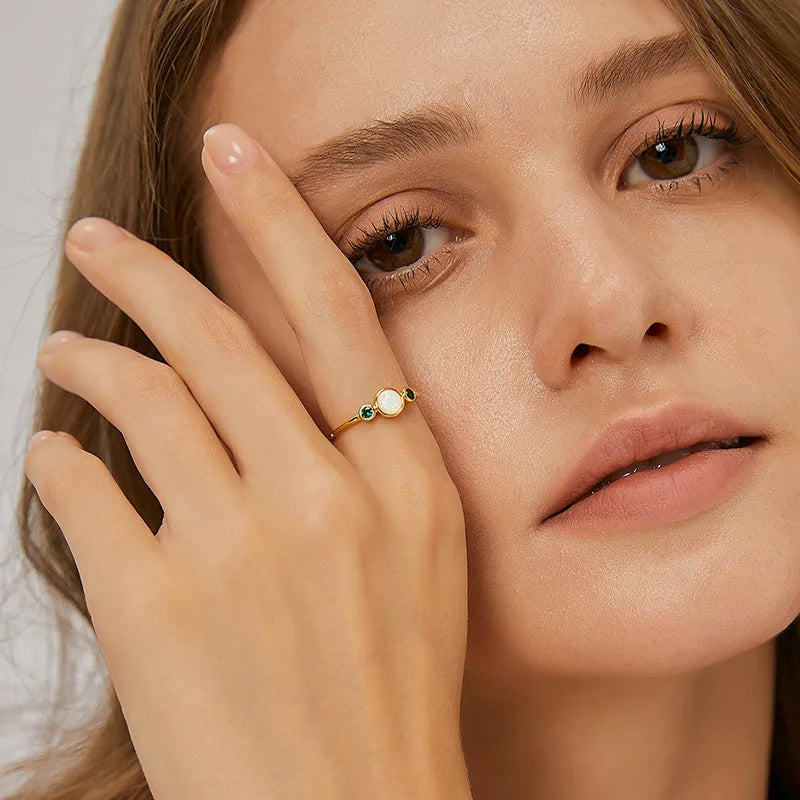 Bague fine plaqué or avec opale blanche irisée au centre et deux zircons verts profonds, au design minimaliste et équilibré, offrant une élégance naturelle et lumineuse.