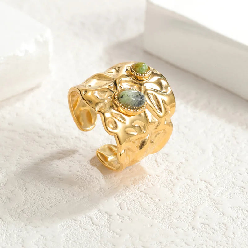 Adoptez une allure brute et précieuse avec cette bague large dorée en acier inoxydable, au design martelé façon métal fondu. Deux pierres naturelles vertes en cabochon viennent illuminer sa structure organique, pour un effet à la fois mystique et résolument moderne.