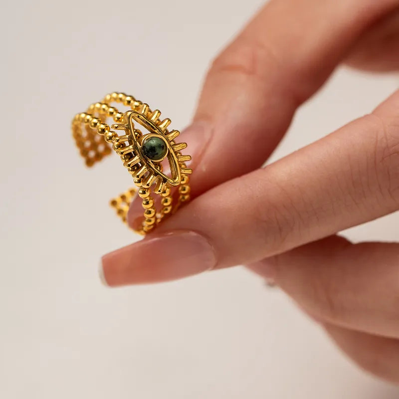 Affichez votre force intérieure avec cette&nbsp;bague œil dorée en acier inoxydable, au design symbolique et structuré. Son motif d’œil protecteur est rehaussé par une pierre ronde verte centrale, entourée de trois rangées de perles dorées texturées. Une pièce forte et mystique à porter au quotidien.