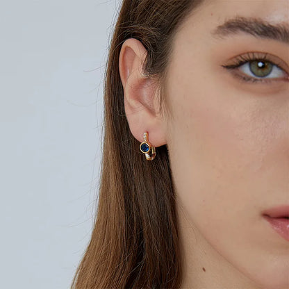Boucles d’oreilles dorées pour femme en plaqué or, ornées d’une pierre bleue facettée effet saphir, d’une lignée de zircons étincelants et de détails en émail blanc. Bijou graphique et sophistiqué au style art déco moderne, idéal pour une tenue habillée ou chic