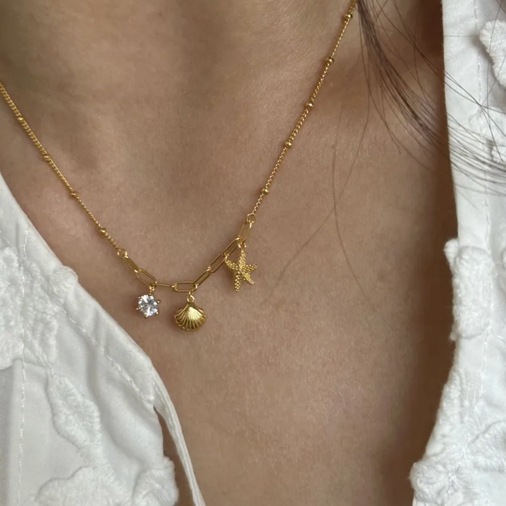 Ce collier doré revisite le thème marin avec élégance. Une chaîne mixte associe maillons larges et perles fines, rehaussée de trois pendentifs évocateurs : une coquille, une étoile de mer et une pierre claire scintillante. Un bijou en acier inoxydable conçu pour résister au temps, tout en évoquant les vacances.

Matériaux : Acier inoxydable, finition couleur or
Dimensions : 40 + 5 cm – poids : 3,5 g
Style : Marin, estival, symbolique