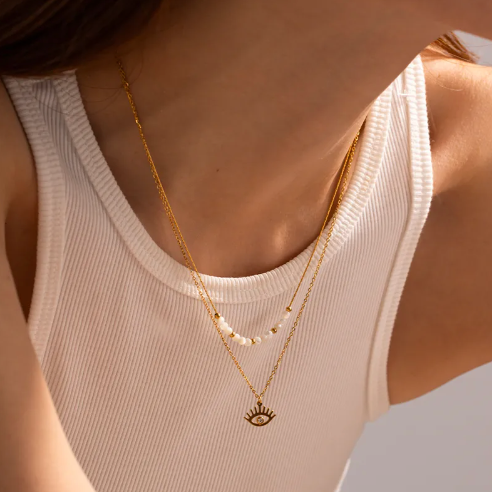 Un collier raffiné à double chaîne, combinant une rangée de perles blanches et dorées avec un pendentif œil protecteur serti d’un strass. Une pièce symbolique et élégante, parfaite pour une touche mystique chic.

Matériau : Acier inoxydable couleur doré, perles synthétiques blanches et dorées, strass central. Résistant à l’eau et hypoallergénique.

Dimensions : Longueur 1ᵉʳ rang 39 + 4 cm – 2ᵉ rang 41 + 4 cm – pendentif œil 1 cm – poids 3,27 g.

Design : Deux chaînes dorées superposées. La première ornée de
