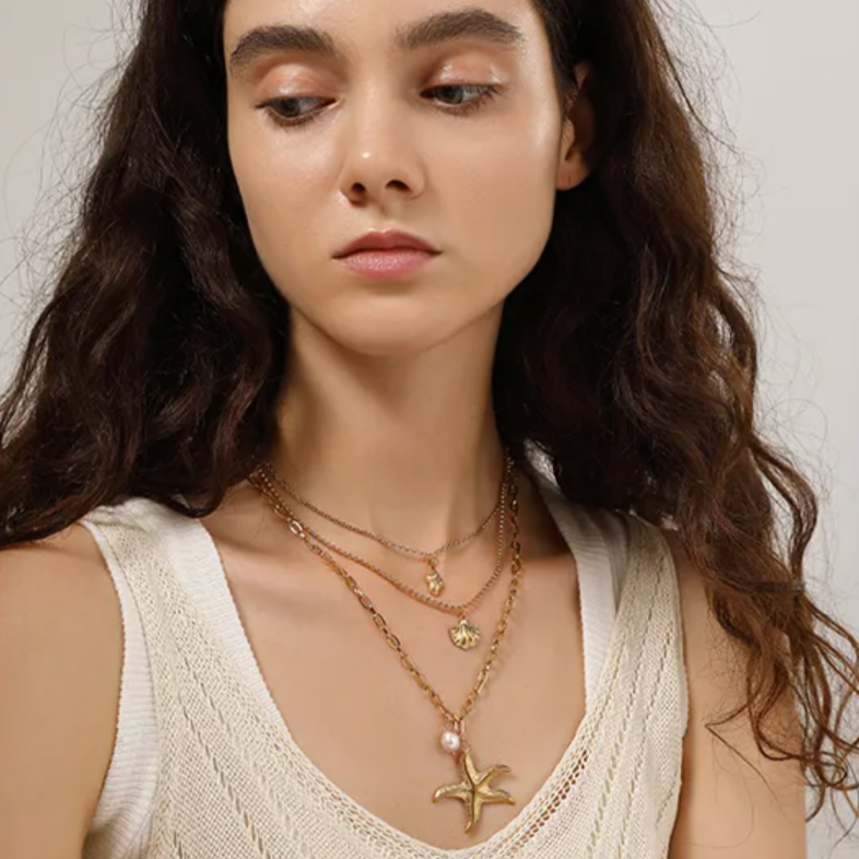 Collier multi-rangs pour femme en acier inoxydable couleur doré, orné de pendentifs coquillage, étoile de mer et perle nacrée. Bijou lumineux et estival au style bohème et marin, parfait pour un look vacances chic