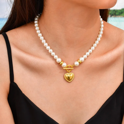 Collier pour femme composé de perles blanches nacrées et de perles dorées, orné d’un pendentif cœur bombé couleur or avec fermoir toggle à l’avant. Bijou romantique et vintage en acier inoxydable, résistant à l’eau et hypoallergénique.
