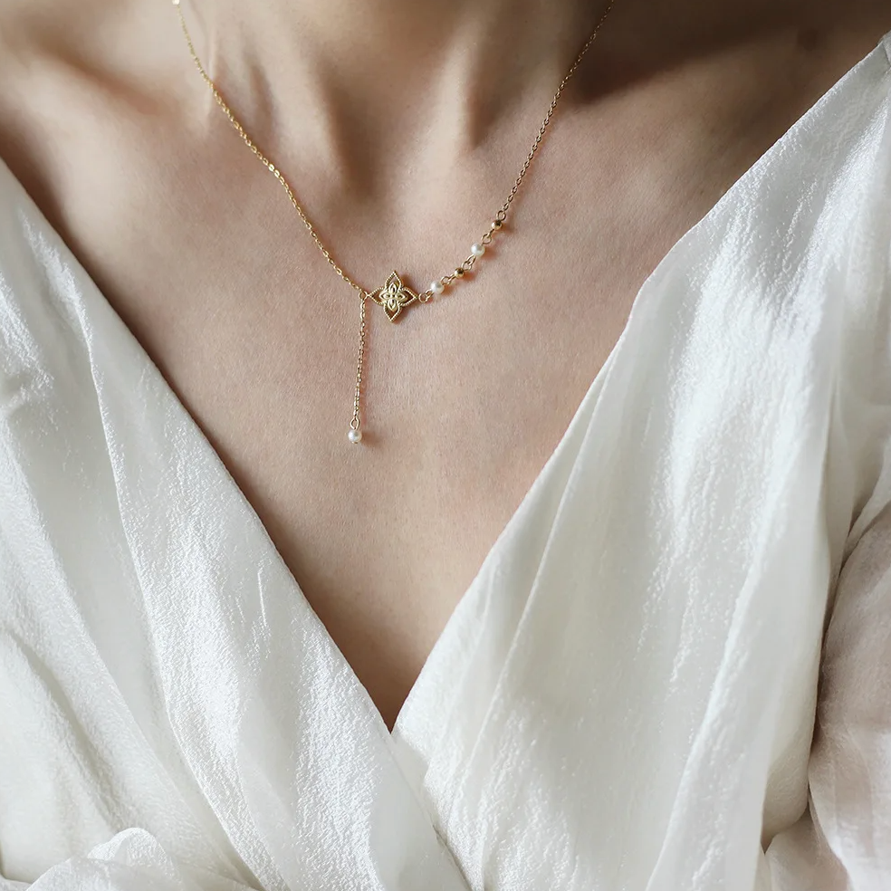 Ce collier doré asymétrique à pendentif fleur dévoile un équilibre subtil entre tradition et modernité. Le motif floral ciselé, suspendu à une fine chaîne prolongée, s'accompagne de petites perles blanches et de sphères dorées, évoquant un bijou raffiné inspiré des arts orientaux.
