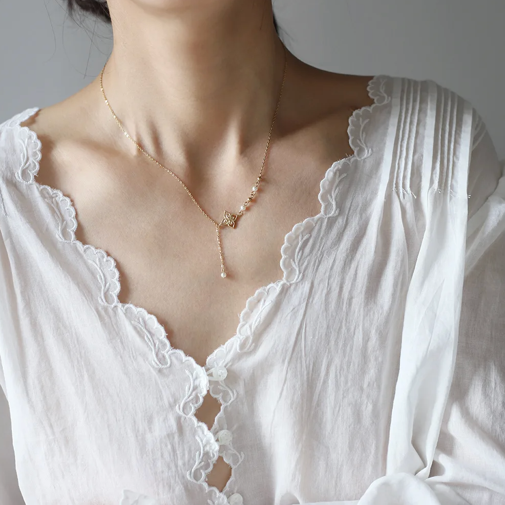Ce collier doré asymétrique à pendentif fleur dévoile un équilibre subtil entre tradition et modernité. Le motif floral ciselé, suspendu à une fine chaîne prolongée, s'accompagne de petites perles blanches et de sphères dorées, évoquant un bijou raffiné inspiré des arts orientaux.