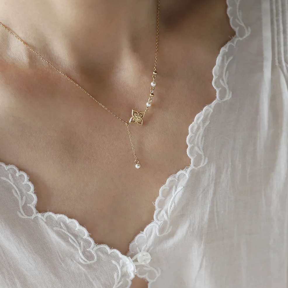 Ce collier doré asymétrique à pendentif fleur dévoile un équilibre subtil entre tradition et modernité. Le motif floral ciselé, suspendu à une fine chaîne prolongée, s'accompagne de petites perles blanches et de sphères dorées, évoquant un bijou raffiné inspiré des arts orientaux.